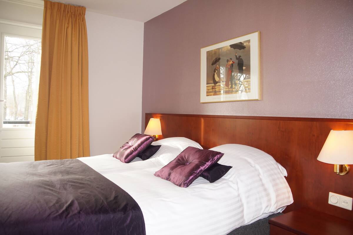A hotel room with a large bed, two bedside lamps, a framed picture on the wall, and a window with curtains.