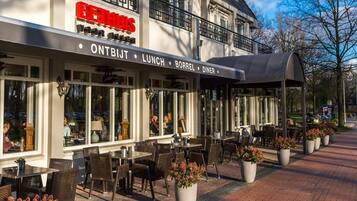 An outdoor cafe with tables and chairs, a covered seating area, and a building with a sign reading "ONTBIJT LUNCH BORREL DINER".