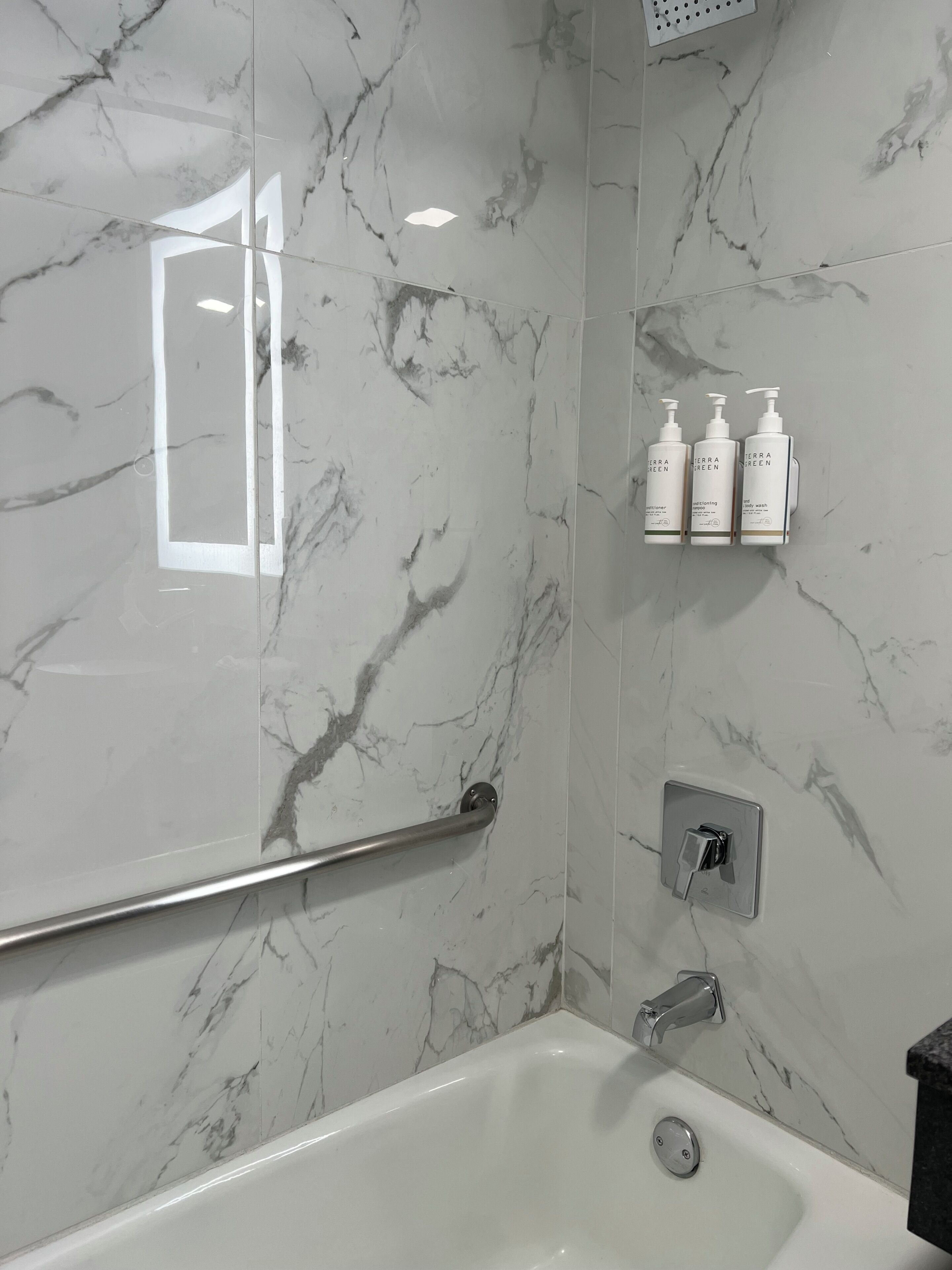 A bathroom with a marble wall, a bathtub, a handrail, a showerhead, and three bottles of Terra Green products.