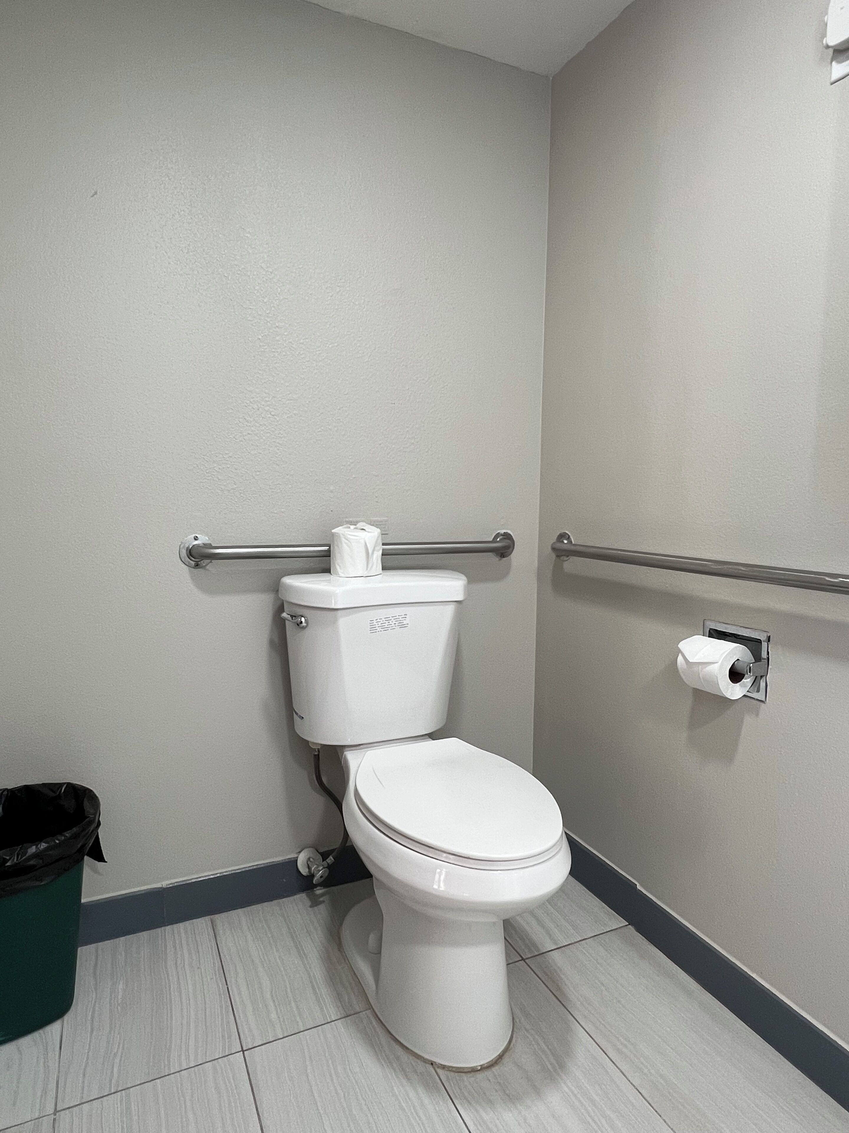 A standard white toilet with a paper towel holder, a grab bar, and a trash bin in a bathroom with light-colored walls and floor tiles.