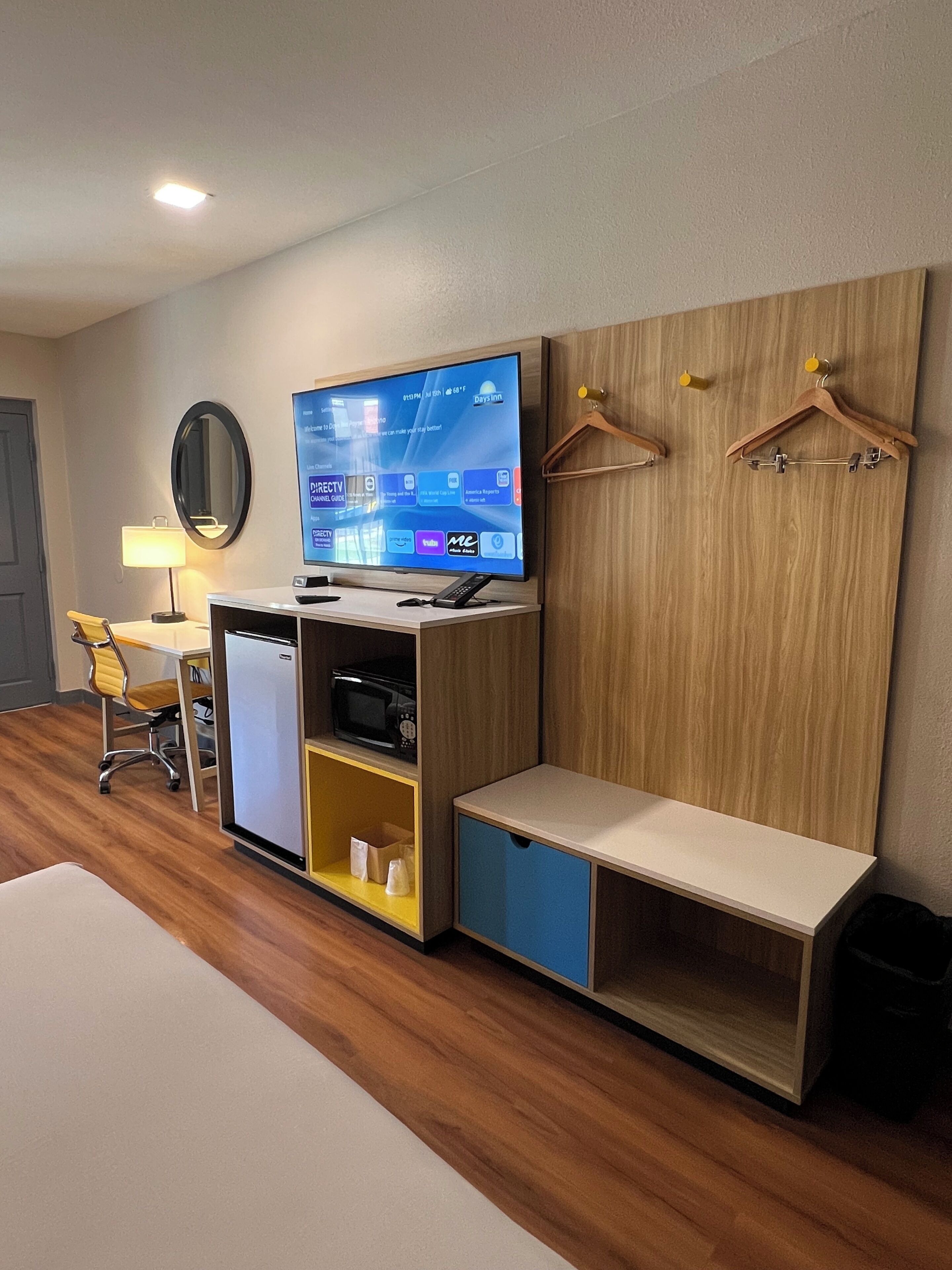 A hotel room with a TV, a desk, a chair, a microwave, and a wardrobe with a bench.