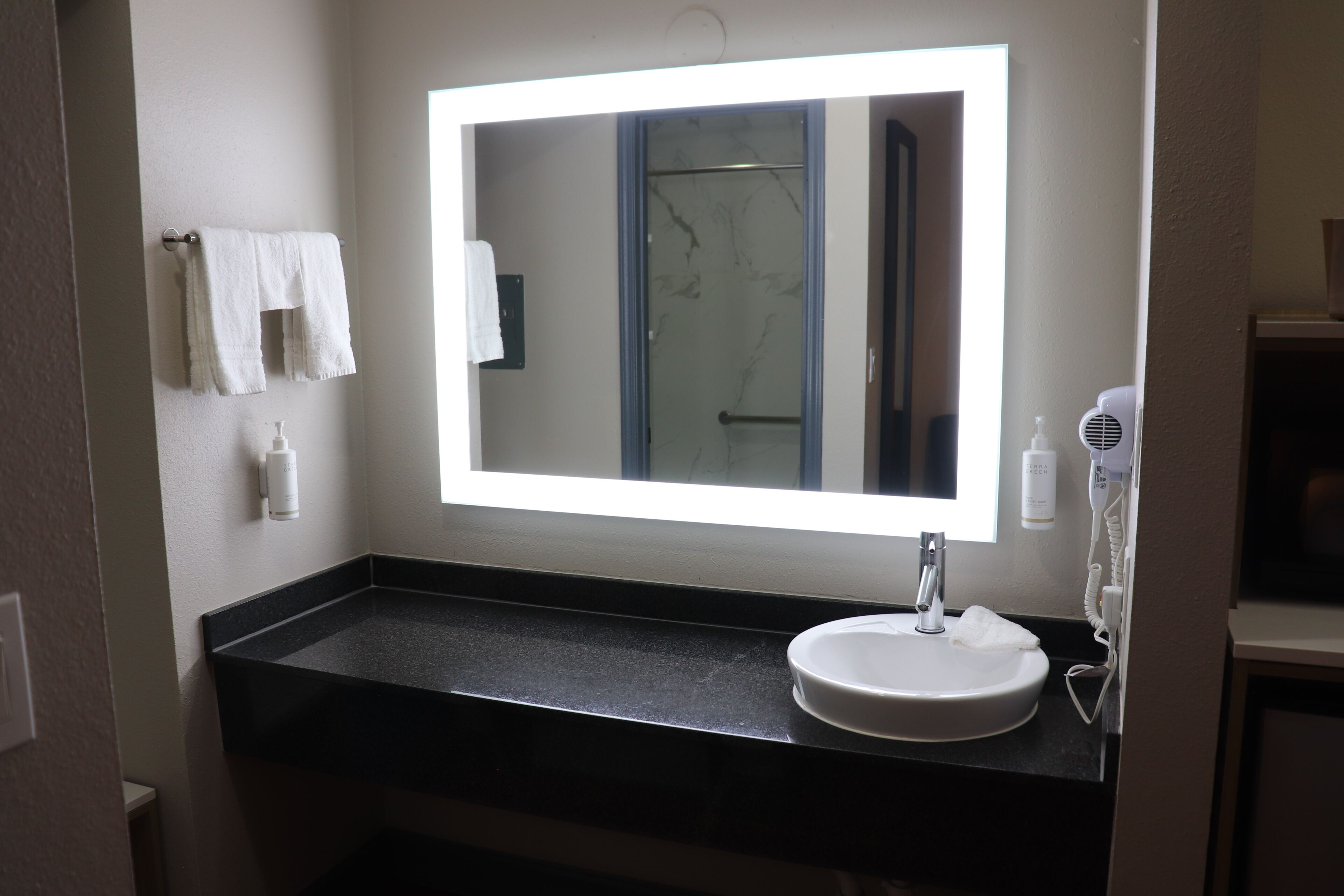 A bathroom with a white sink, a mirror with a light, a towel rack, and a handheld hair dryer.