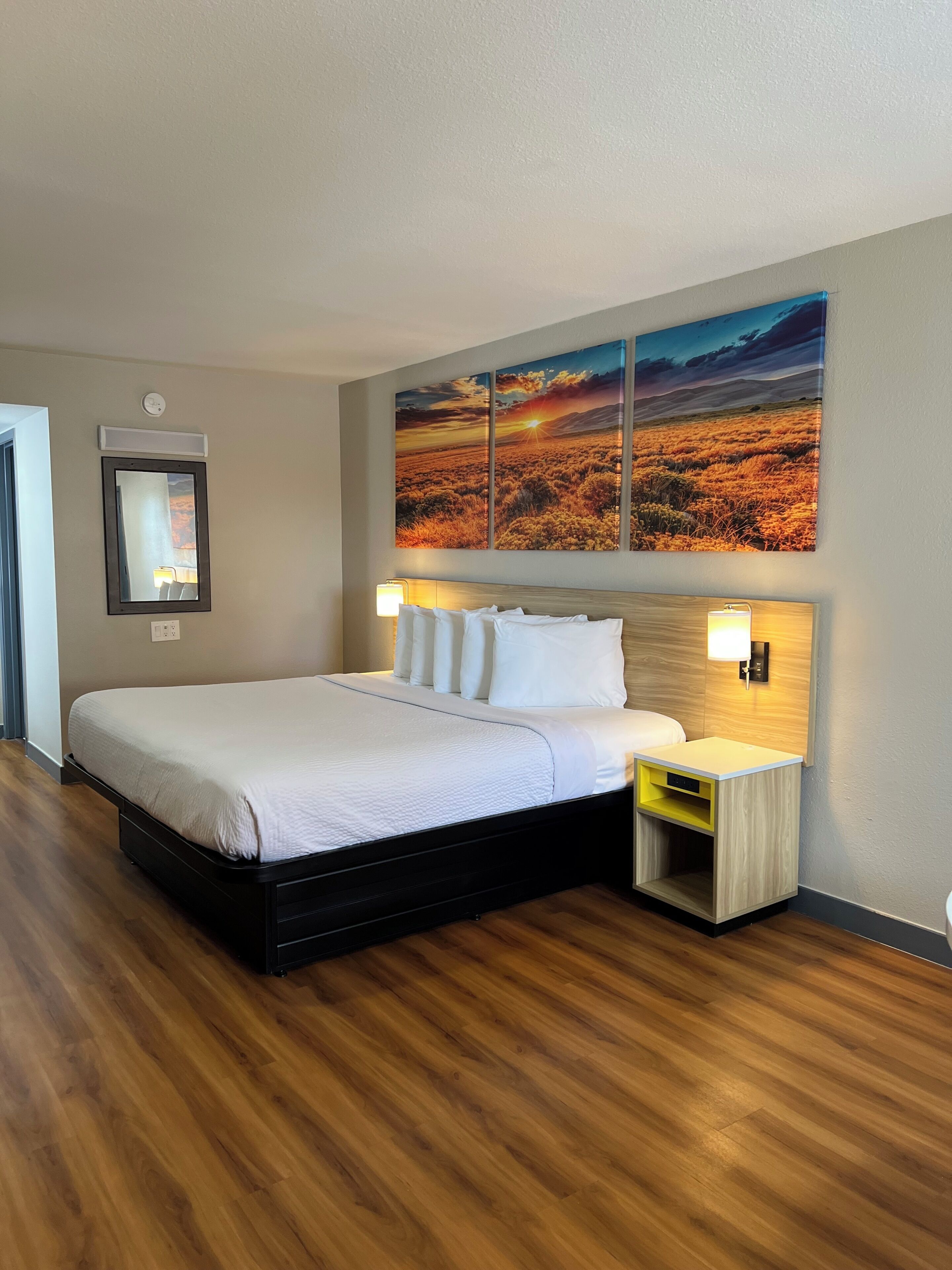 Habitación de hotel con una cama grande, una mesita de noche con lámpara, un espejo montado en la pared y una pintura de paisaje con atardecer.