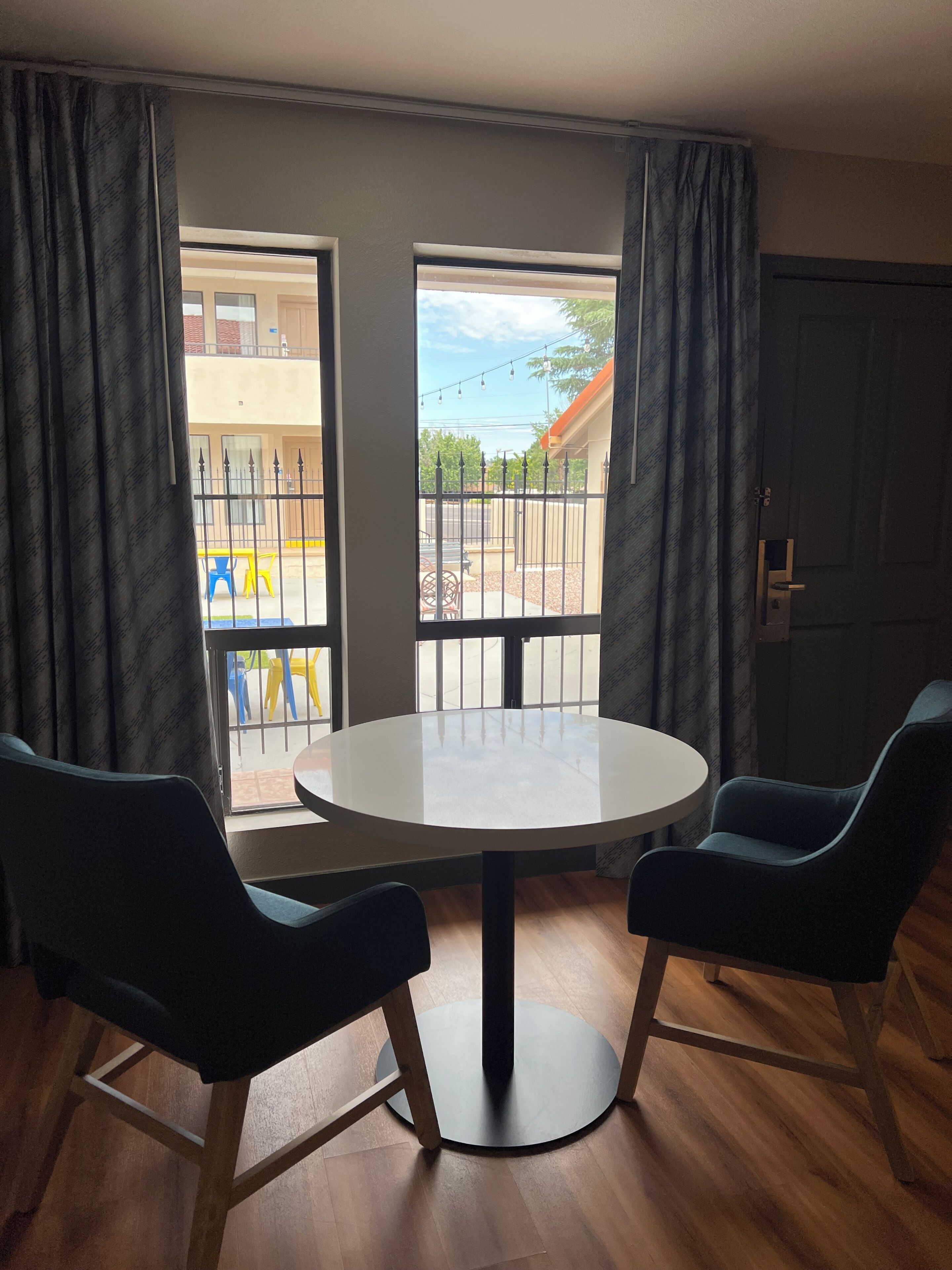 A room with a round table and two chairs, a door, and a view through a window with curtains.