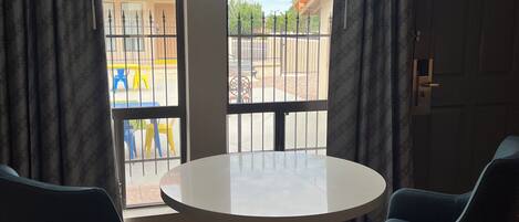 A room with a round table and two chairs, a door, and a view through a window with curtains.