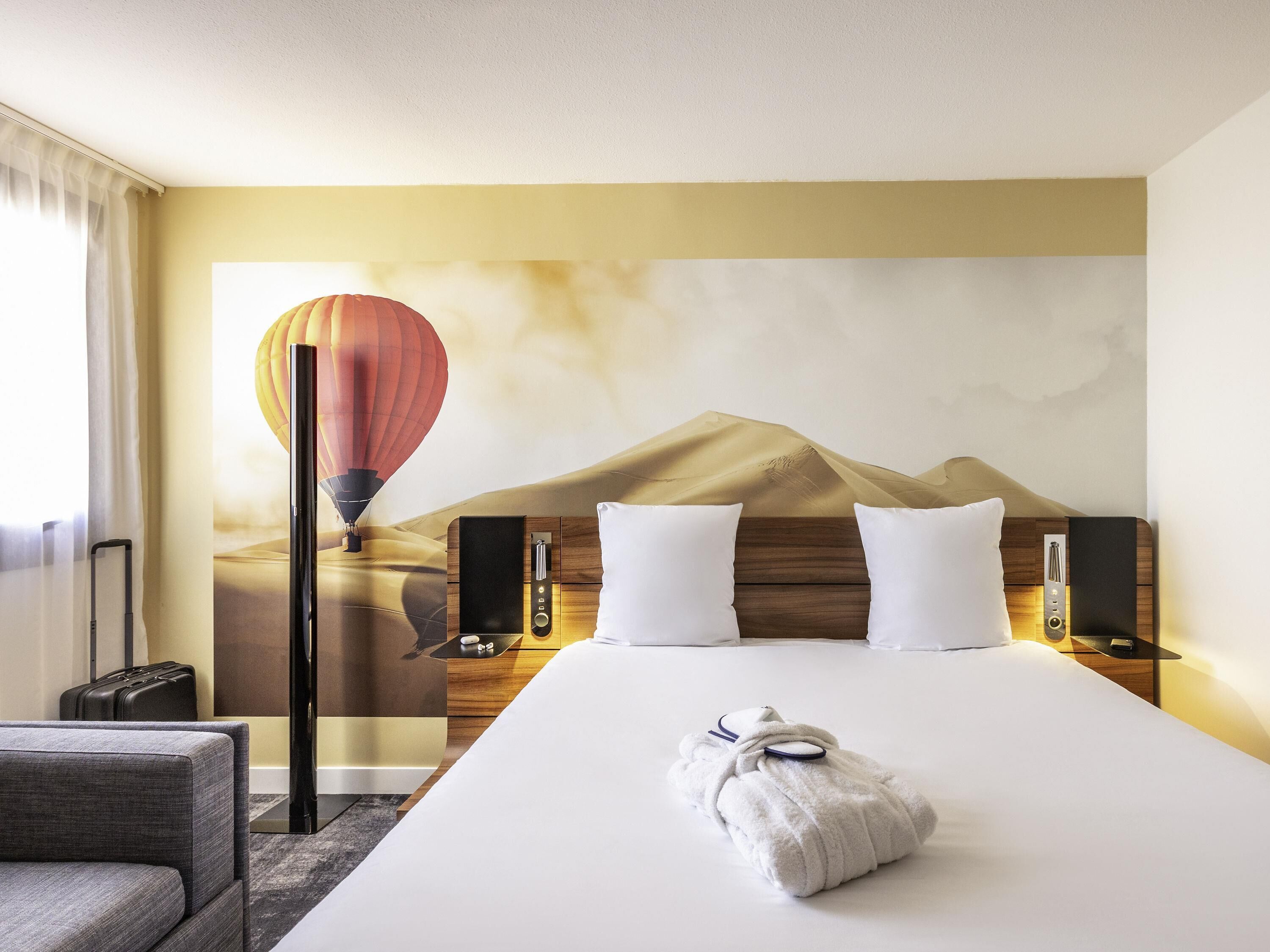 A hotel room with a bed, pillows, a nightstand, a lamp, a hot air balloon artwork, and a suitcase.