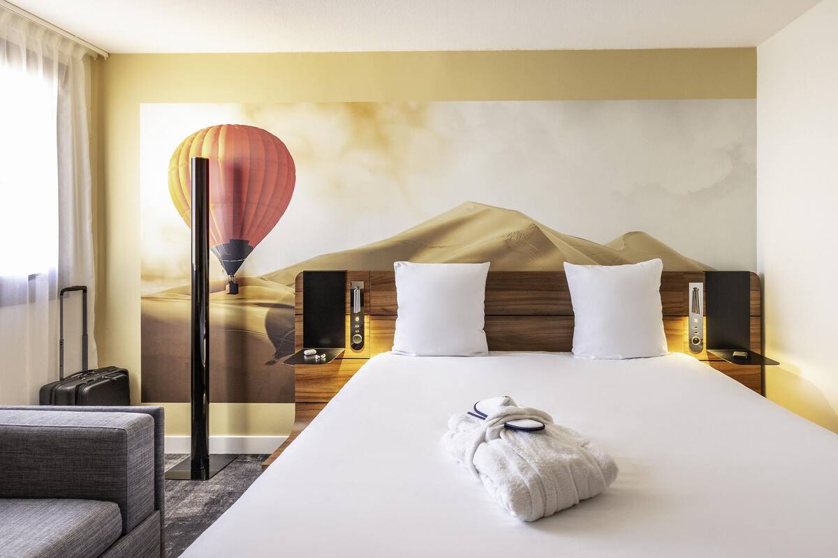 Ein Hotelzimmer mit Bett, Kissen, Nachttisch, Lampe und einem Wandbild mit einer Heißluftballon-Szene. Ein Koffer steht ebenfalls im Raum.