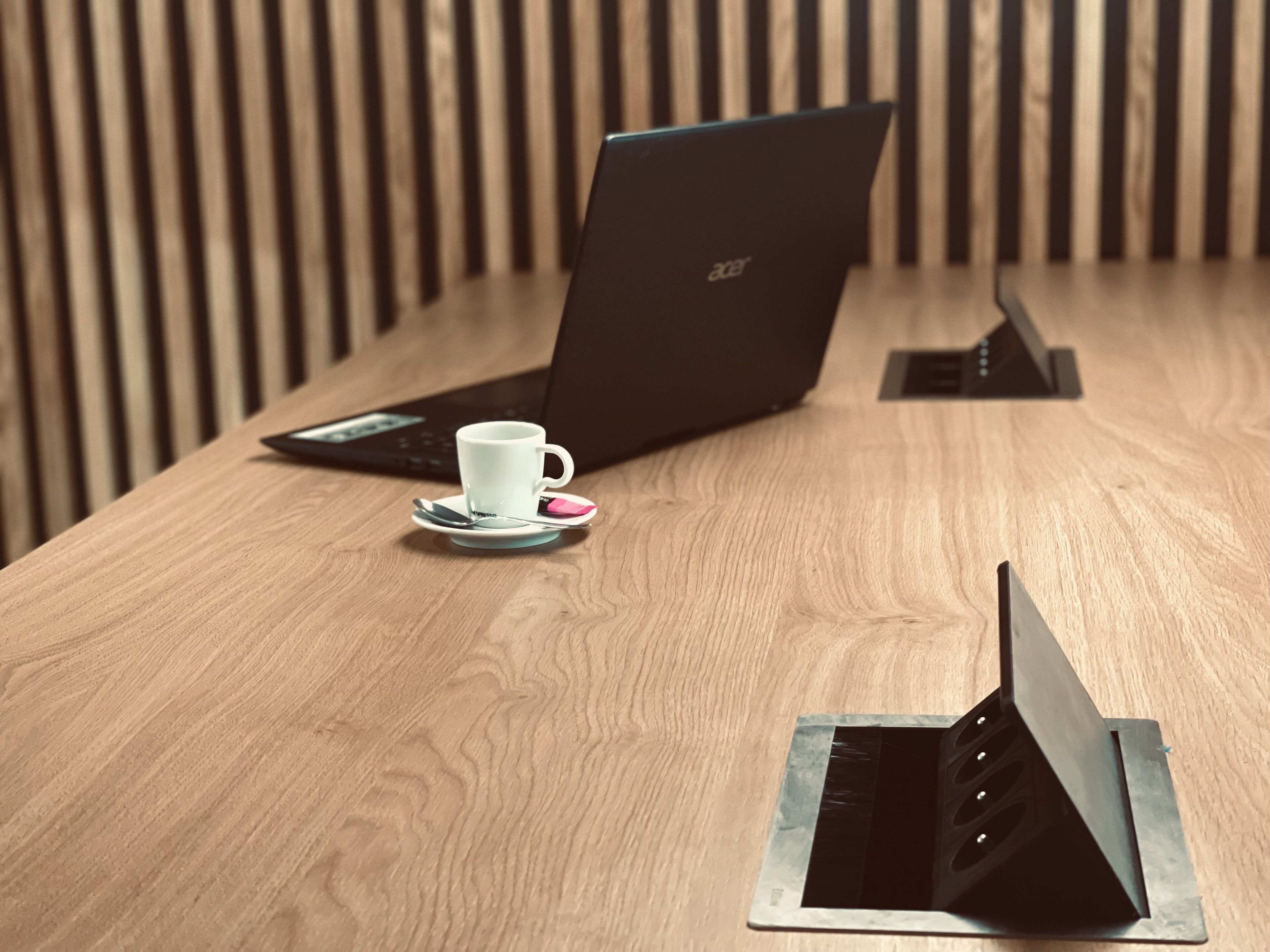 Una mesa de madera con una computadora portátil, una taza blanca y un teléfono inteligente en un soporte.