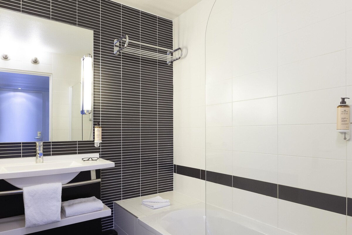 A bathroom with a white sink, a mirror, a towel rack, a shower, and a hand sanitizer dispenser.