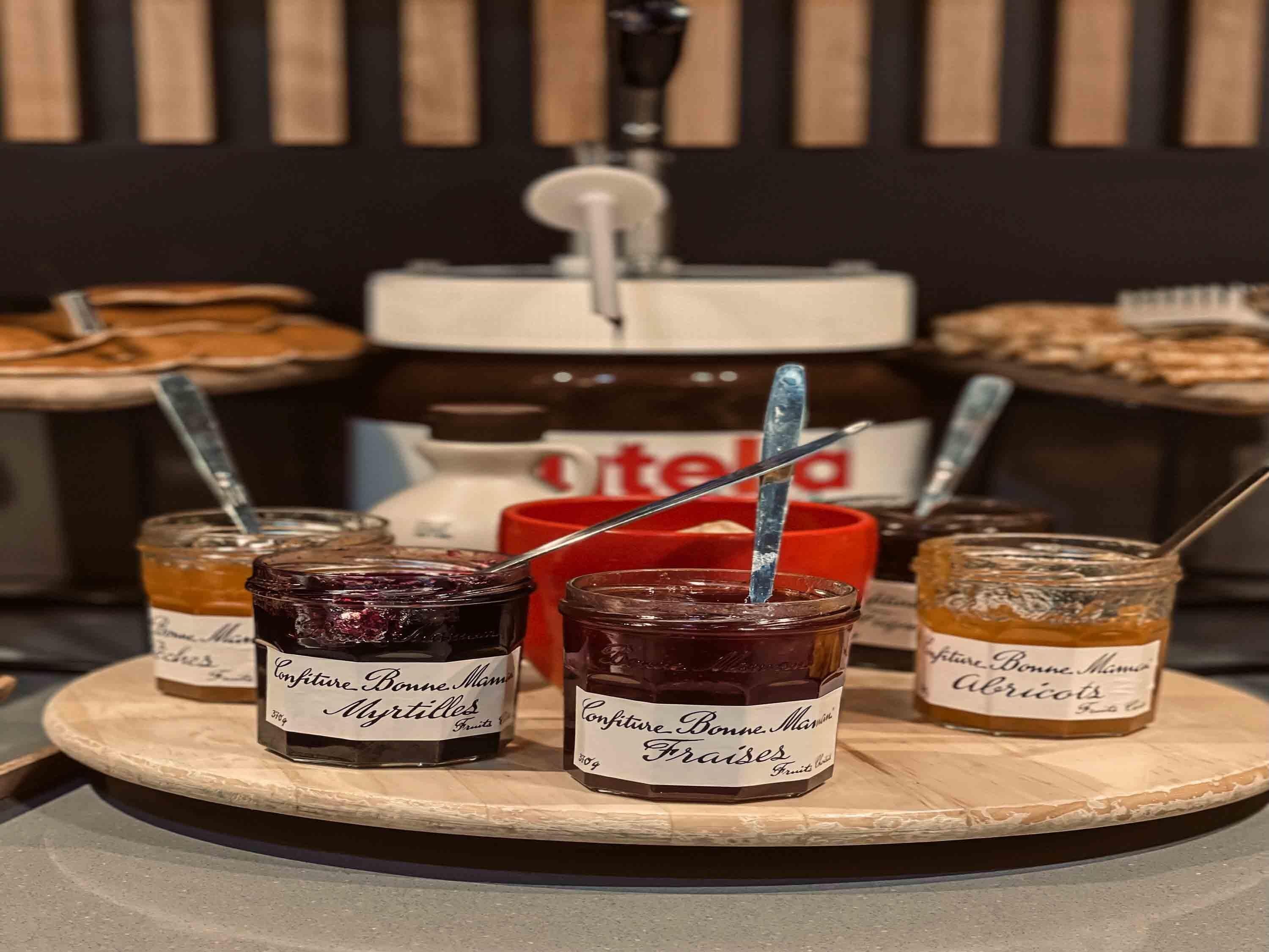 Gläser mit Fruchtkonfitüre auf einem Holzboden, dazu ein Nutella-Glas und im Hintergrund ein roter Behälter.