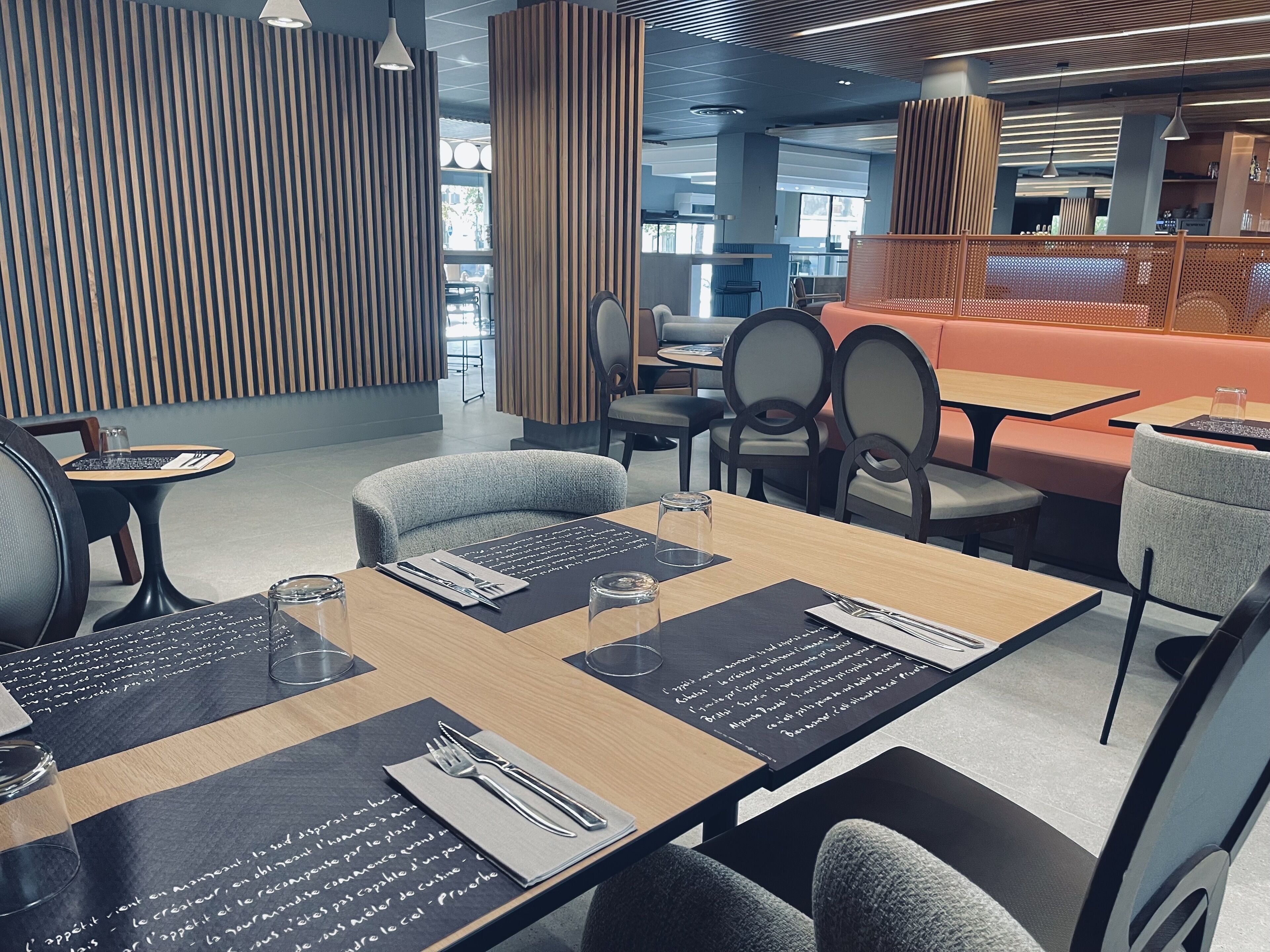 Un restaurant moderne avec des murs lambrés en bois, des sets de table noirs ornés de motifs et un mélange de sièges aux couleurs gris et orange.