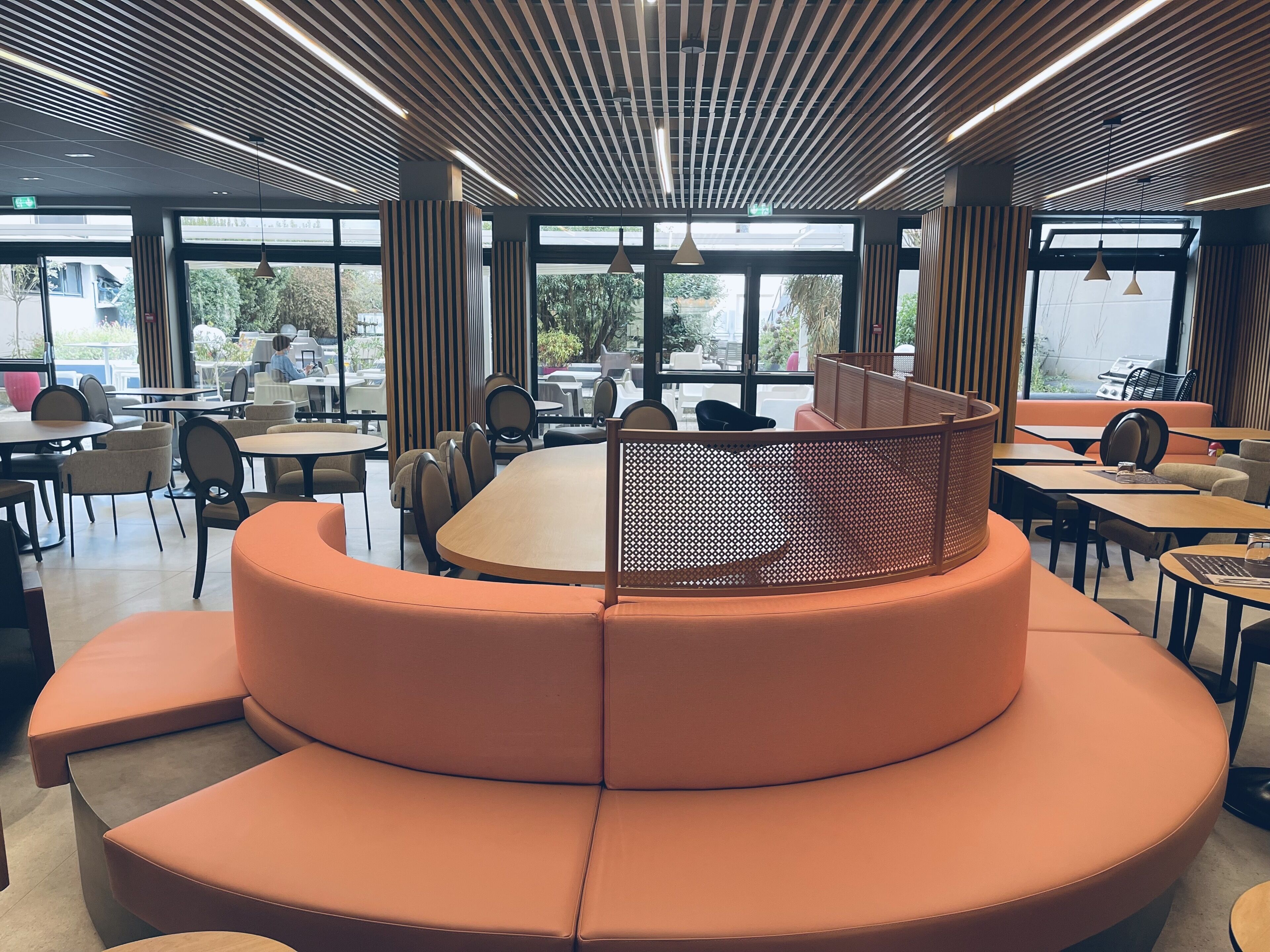 A modern cafe with a central seating area featuring a curved orange sofa, surrounded by tables and chairs. Large windows provide natural light and views of the outdoors.