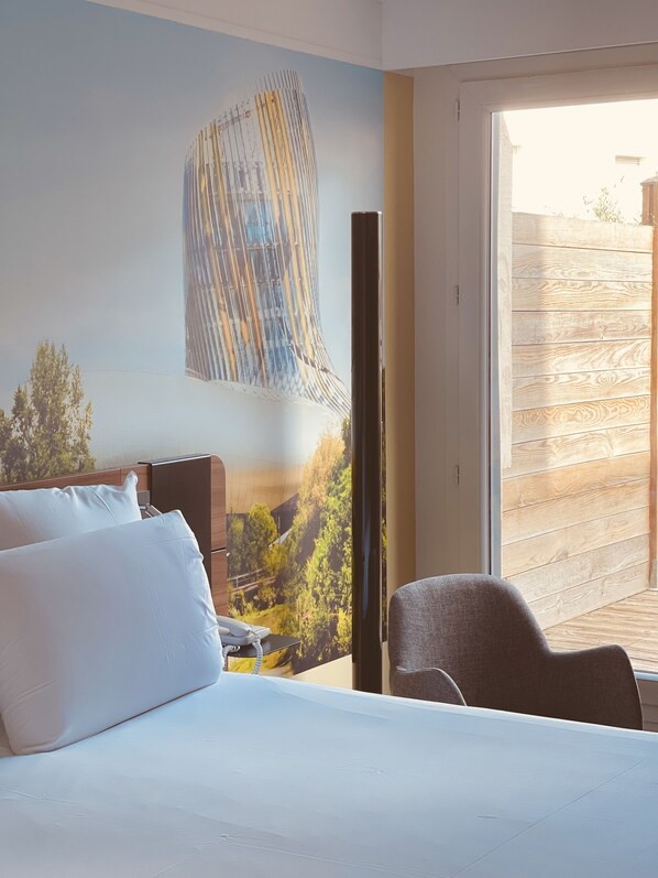 Une chambre d’hôtel avec un lit, une chaise et une grande fenêtre donnant sur un immeuble et des arbres.