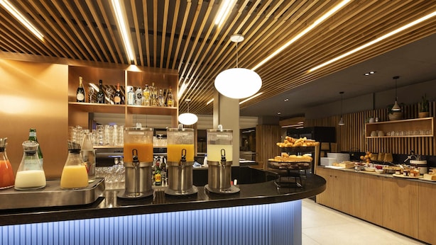 A bar area with multiple beverage dispensers, a counter with food items, and a wooden ceiling with linear lighting. - Novotel Bordeaux Centre Gare Saint Jean (Bordeaux)