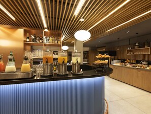 A bar area with multiple beverage dispensers, a counter with food items, and a wooden ceiling with linear lighting. - Novotel Bordeaux Centre Gare Saint Jean (Bordeaux)