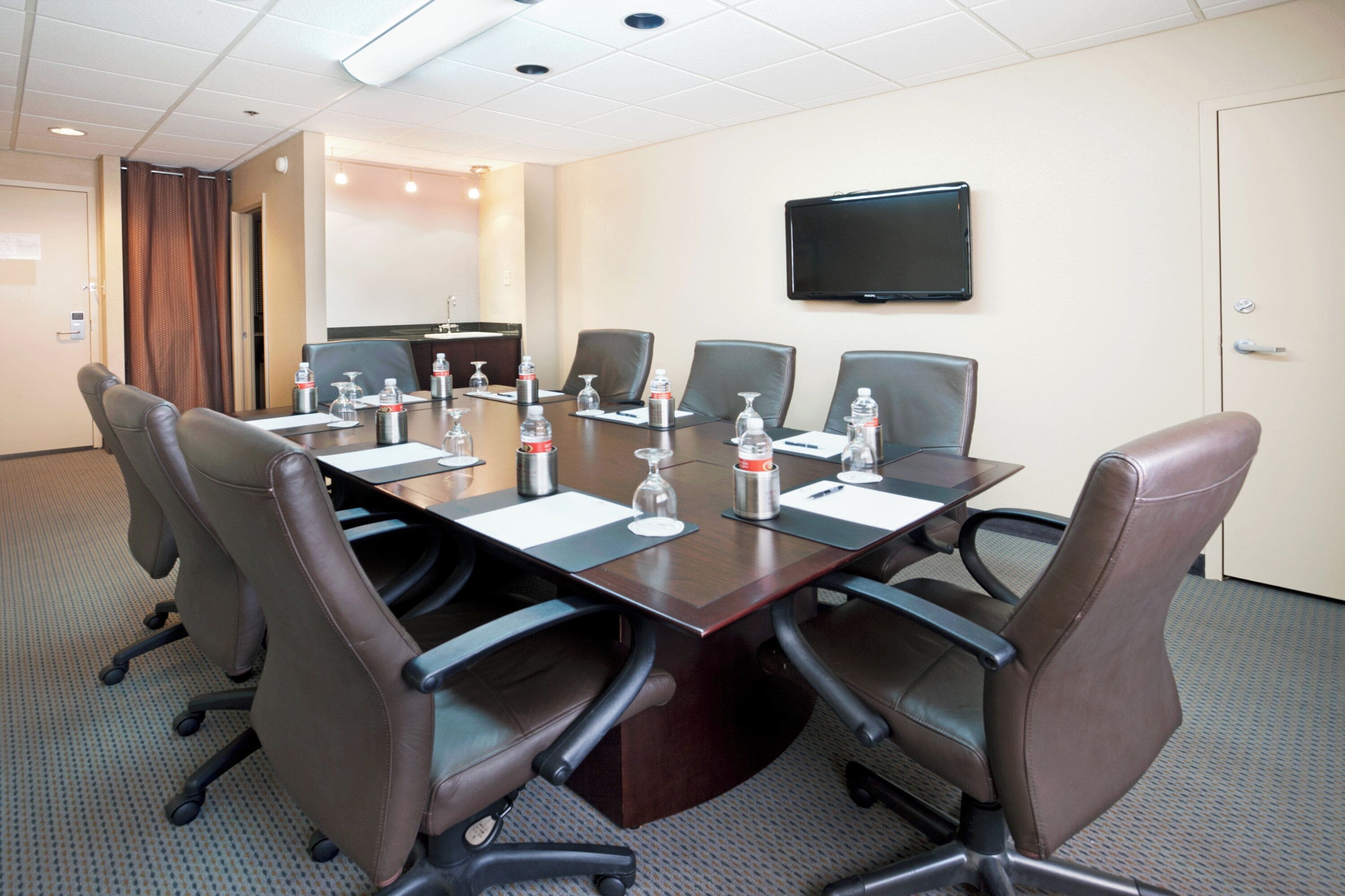 A conference room with a long table, six chairs, water bottles, and a flat-screen TV on the wall.