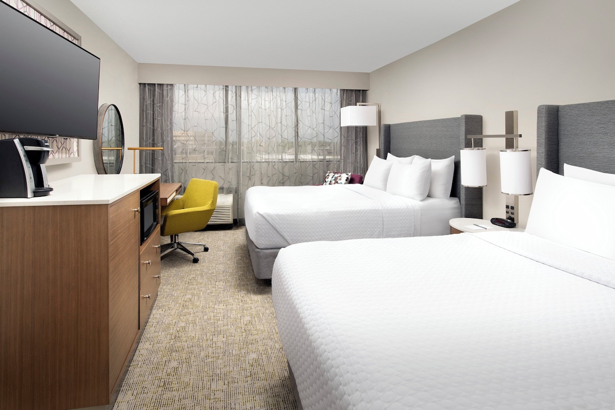 A hotel room with two beds, a desk with a chair, a television, and a window with curtains.
