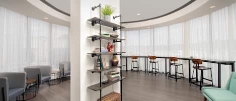 A modern interior with a central shelving unit, a seating area with chairs and a table, and a large window with sheer curtains.