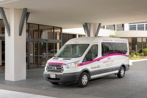 Uma van branca com a marcação 'CROWNE PLAZA HOTELS & RESORTS' estacionada em frente a um edifício com entrada de vidro.
