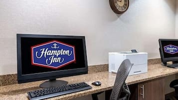 A Hampton Inn reception desk with a computer monitor displaying the hotel's name, a clock on the wall, and a printer.