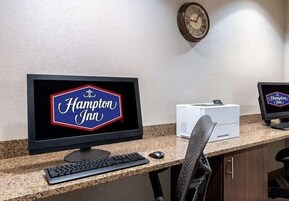 Mostrador de recepción de Hampton Inn, con un monitor que muestra el nombre del hotel, un reloj en la pared y una impresora.