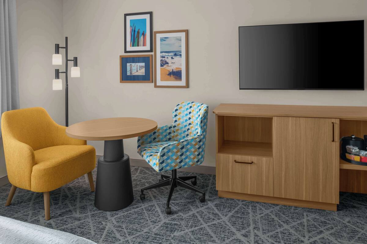 Una habitación con un sillón amarillo, una silla de escritorio estampada, una mesa redonda, un televisor y un mueble de madera.