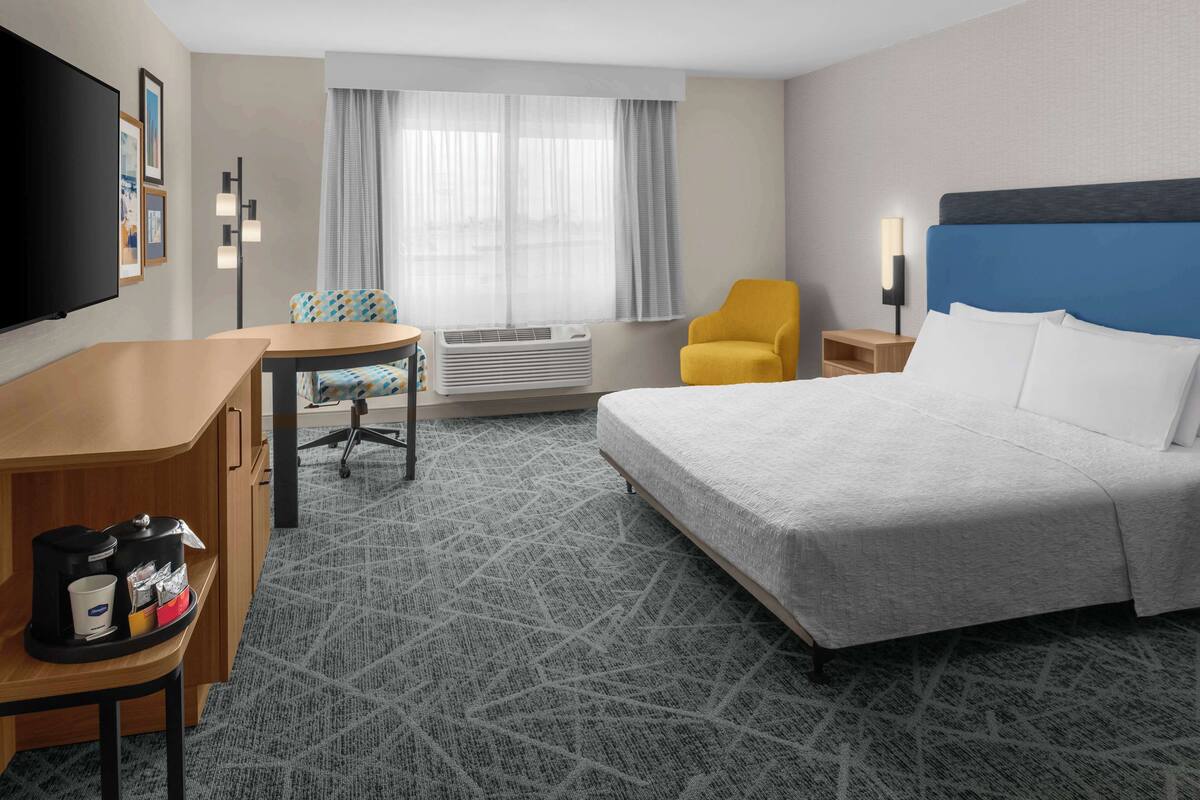 Habitación de hotel con cama, escritorio con cafetera, silla, televisor y ventana con cortinas.