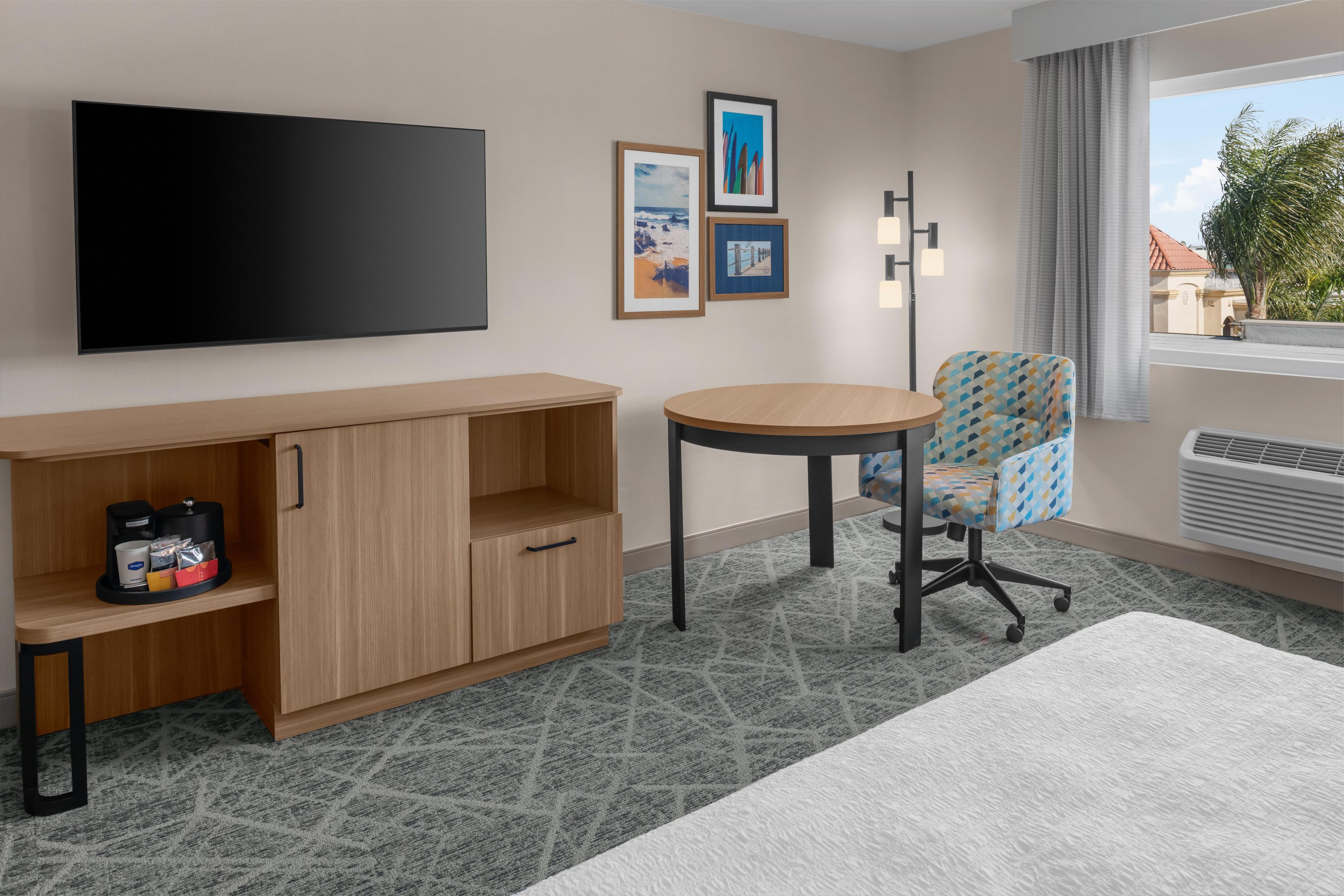 Une chambre d’hôtel équipée d’un téléviseur à écran plat, d’un meuble en bois contenant une machine à café, d’une table ronde, d’un fauteuil à motifs et d’une fenêtre donnant sur des immeubles et des palmiers.