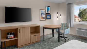 Une chambre d’hôtel équipée d’un téléviseur à écran plat, d’un meuble en bois contenant une machine à café, d’une table ronde, d’un fauteuil à motifs et d’une fenêtre donnant sur des immeubles et des palmiers.