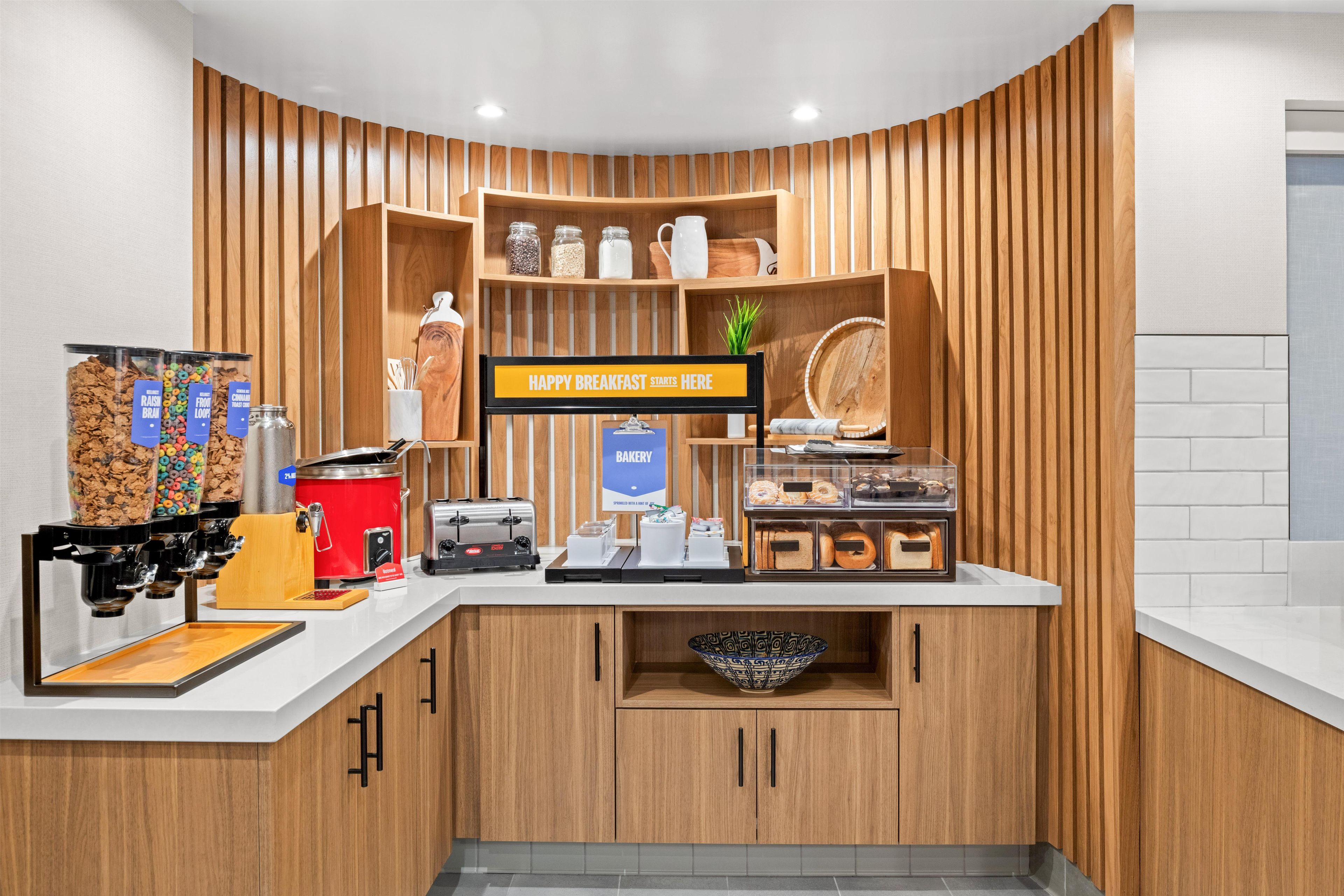 A breakfast area with a sign that reads 'HAPPY BREAKFAST STARTS HERE,' a coffee machine, a toaster, and various food items on display.