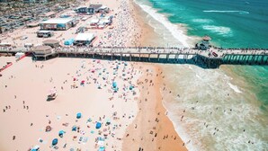 Vista aérea de una playa con un muelle, numerosos paraguas y gente.