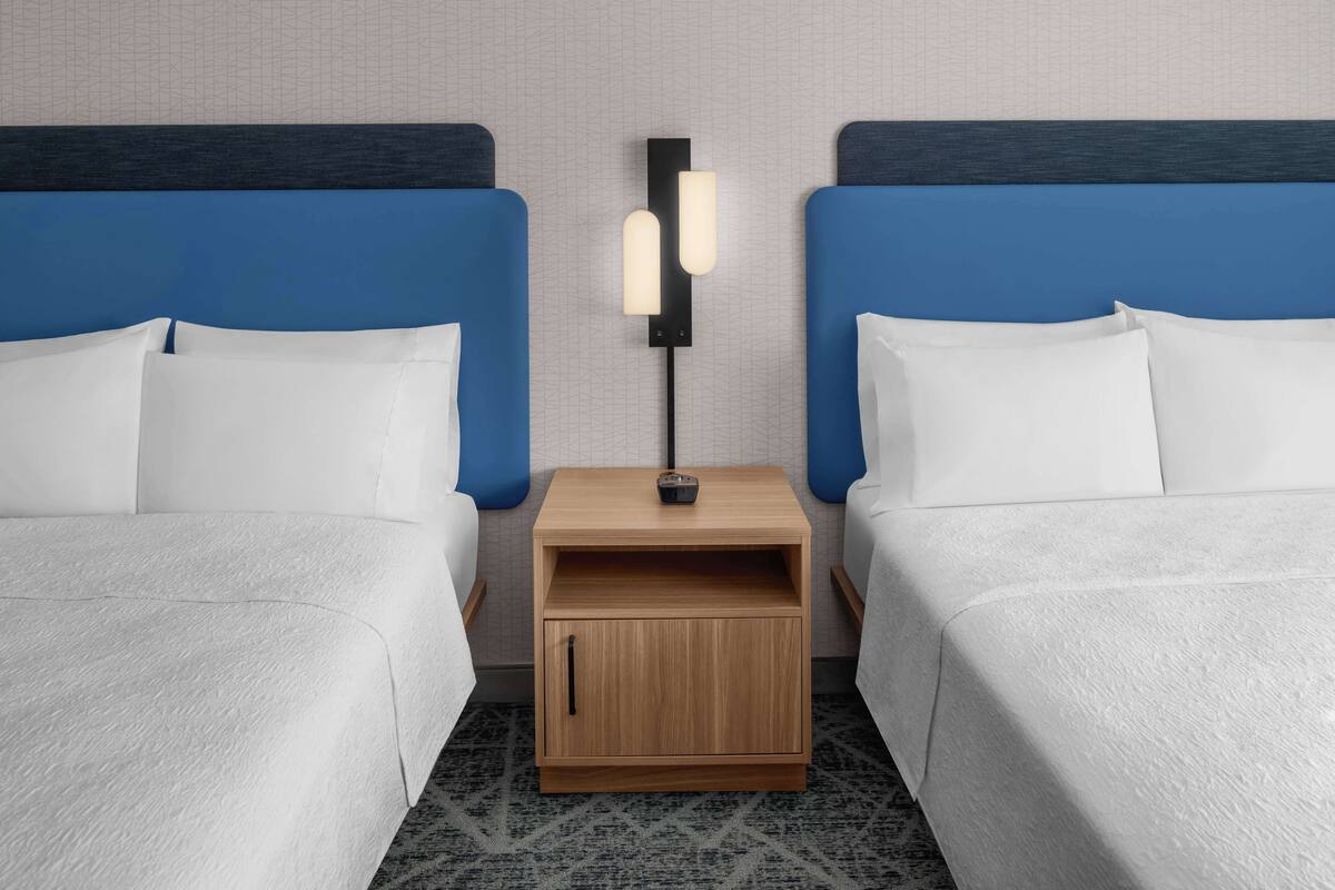 Habitación de hotel con dos camas, cada una con cabecero azul y ropa de cama blanca. Entre las camas, una mesita de noche de madera con una lámpara.