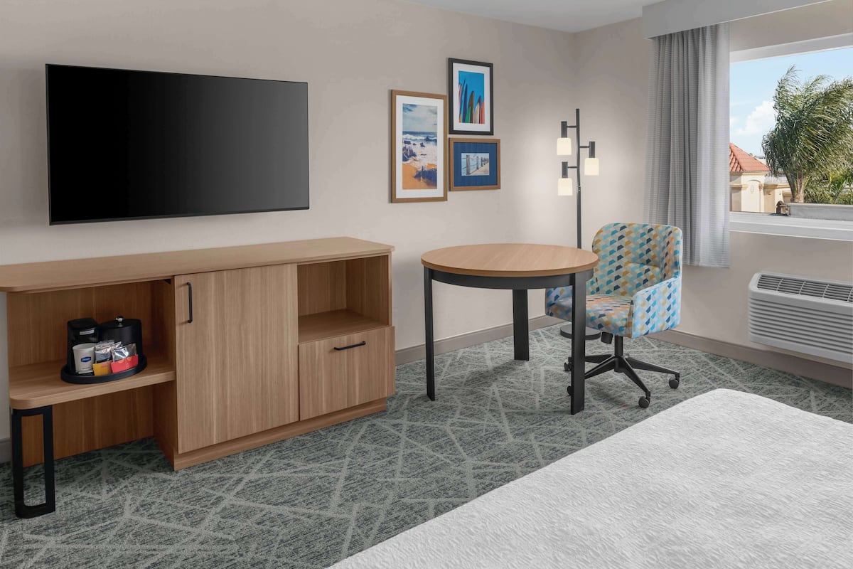 Habitación de hotel con televisor de pantalla plana, un mueble de madera, una mesa redonda, una silla estampada y vistas al exterior.