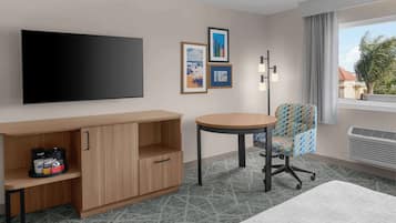A hotel room with a flat-screen TV, a wooden cabinet, a round table, a patterned chair, and a view of the outside.