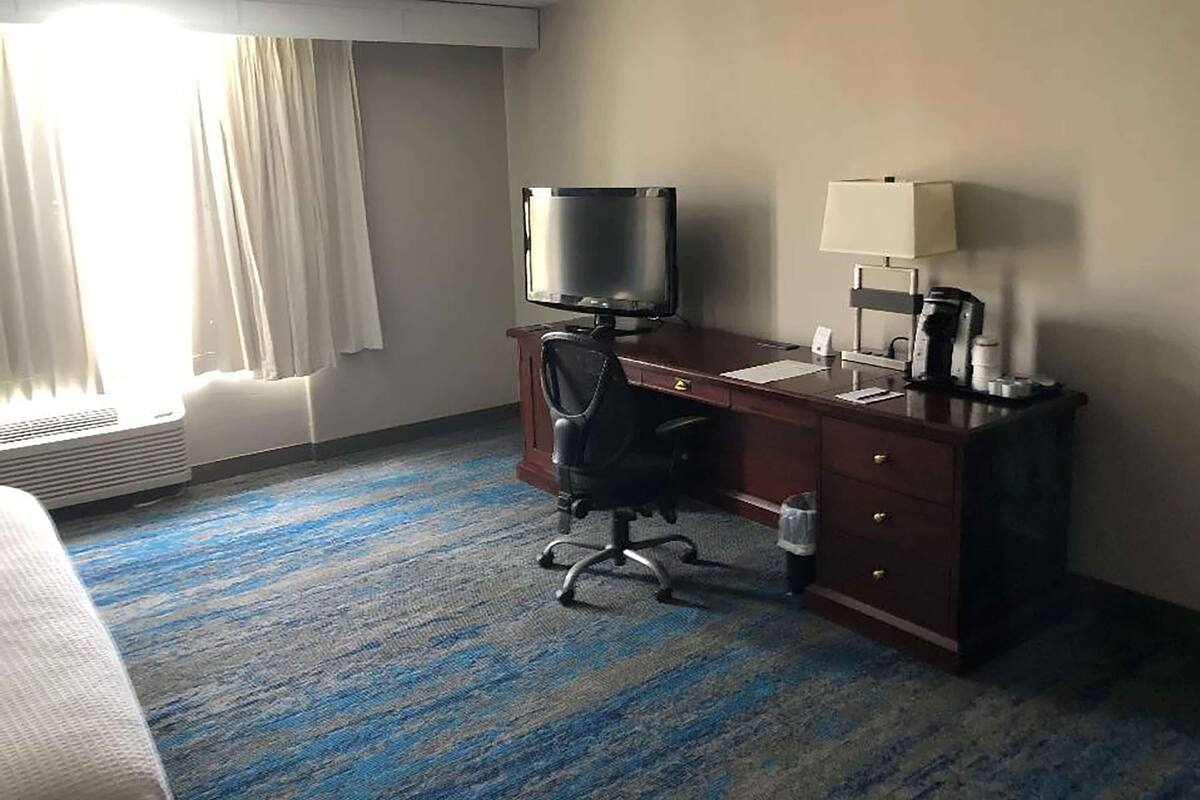 Habitación de hotel con una cama, un escritorio con computadora y lámpara, una silla y un ventanal con cortinas.
