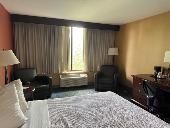 A hotel room with a bed, two chairs, a desk, a lamp, and a window with curtains.