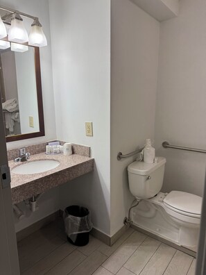 A bathroom with a granite countertop sink, a mirror, a toilet with a grab bar, and a trash can. - Ramada by Wyndham Cleveland Independence (Independence)