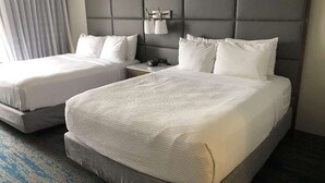 A hotel room with two beds, a grey padded headboard, white linens, and a nightstand with a lamp.