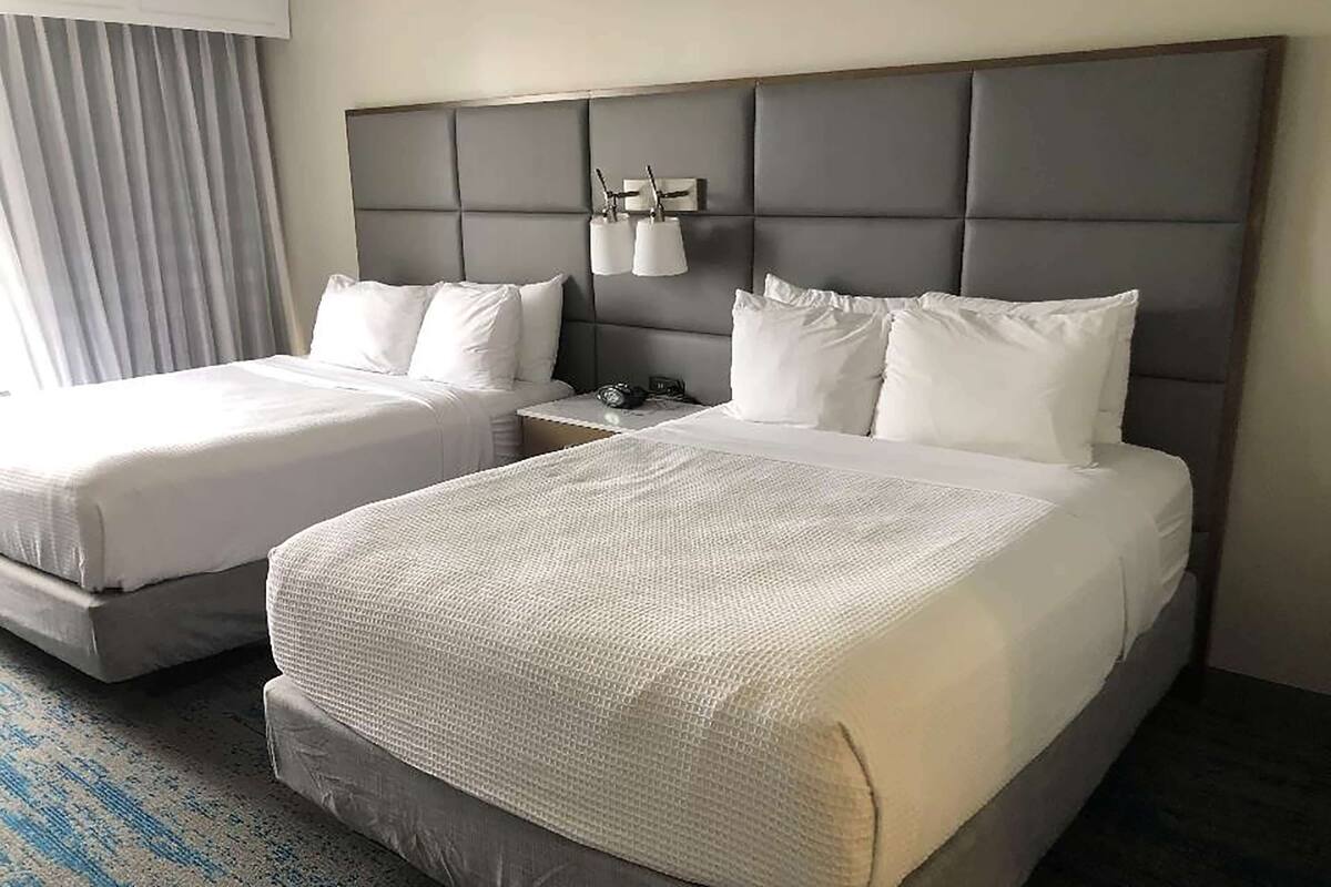 Habitación de hotel con dos camas, un cabecero acolchado gris, ropa de cama blanca y una mesita de noche con lámpara.