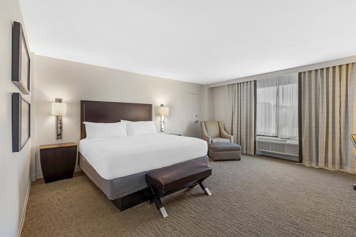 Une chambre d’hôtel avec un grand lit, des lampes de chevet, une chaise et une fenêtre avec des rideaux.
