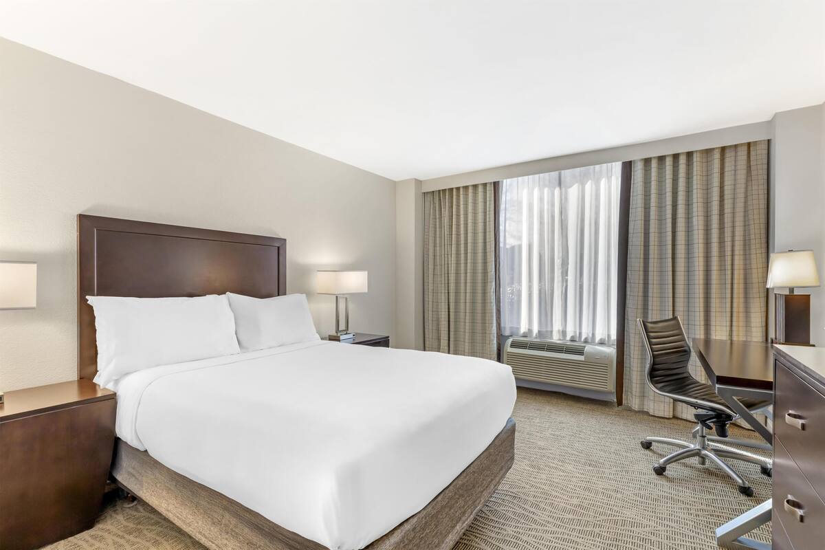 Une chambre d’hôtel avec un grand lit, un bureau avec une chaise, une lampe et une fenêtre avec des rideaux.