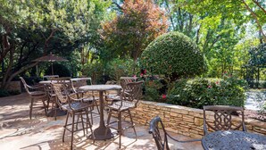 Une terrasse extérieure avec un sol en pierre, des tables et des chaises en métal, entourée d’une végétation luxuriante et d’un mur en pierre.