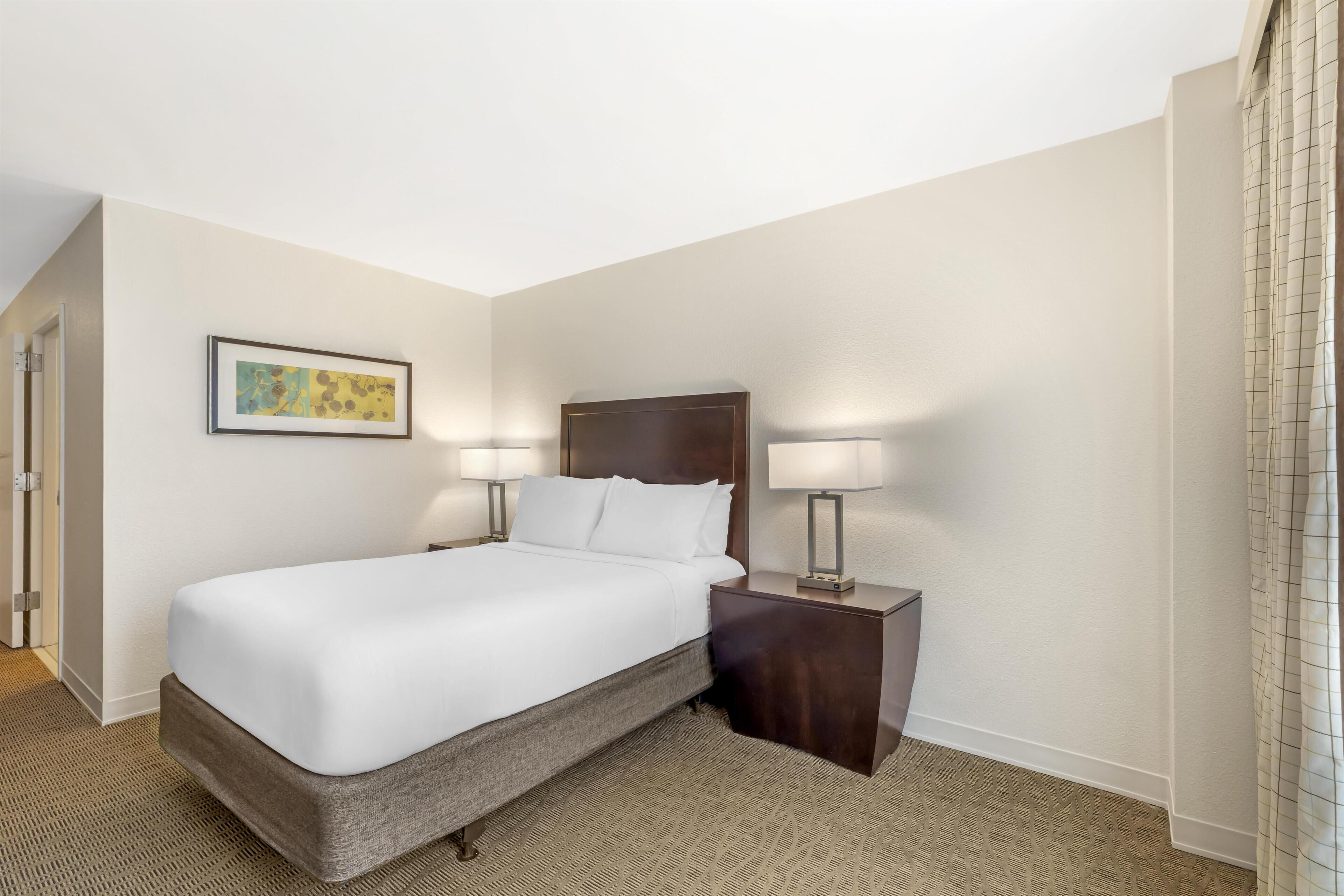 A hotel room with a large bed, two bedside lamps, a framed picture on the wall, and a carpeted floor.