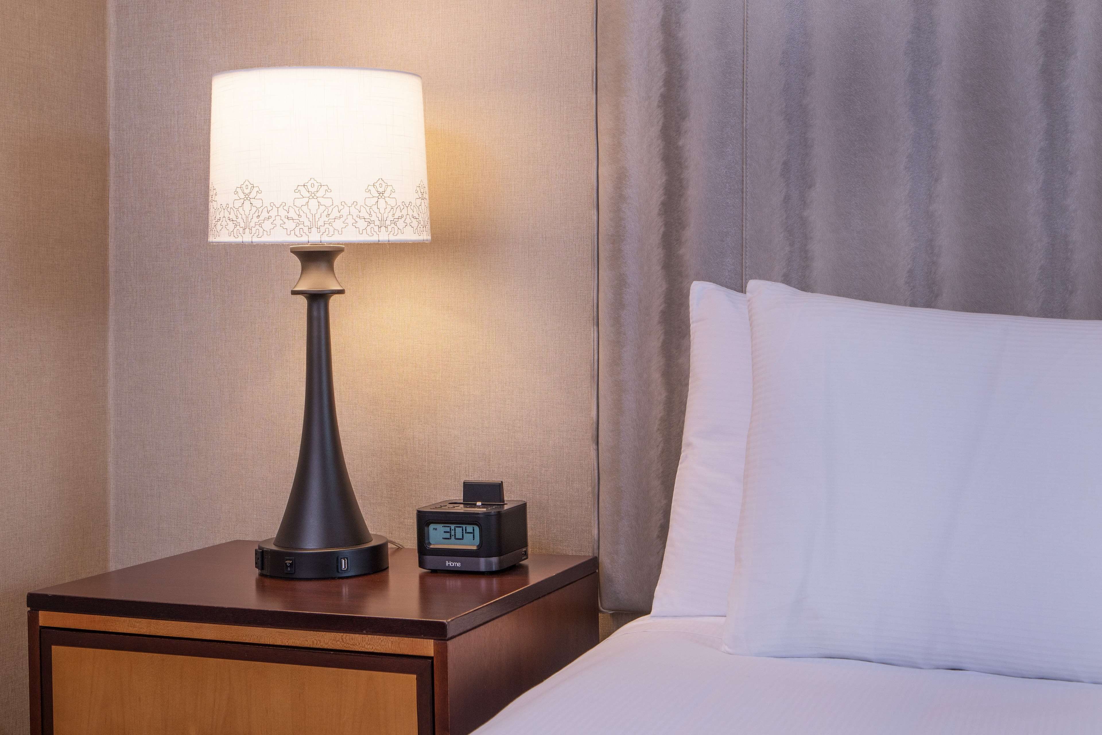 A neatly made bed with white linens, a bedside table with a lamp and a digital clock, and a curtain in the background.