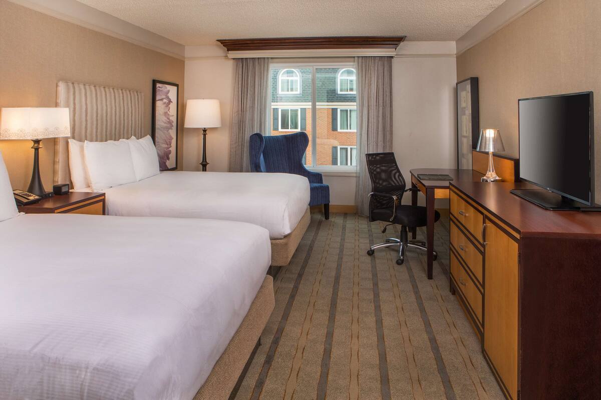 Een hotelkamer met twee bedden, een bureau, een stoel, een televisie en een raam met uitzicht op gebouwen.