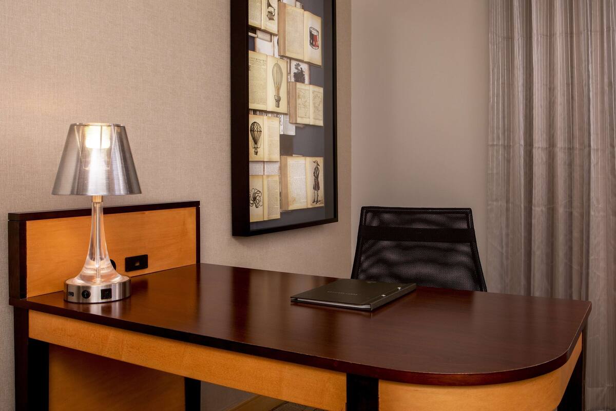 Een hotelkamer met een houten bureau, een lamp, een lijst met collages aan de muur en een zwarte stoel.