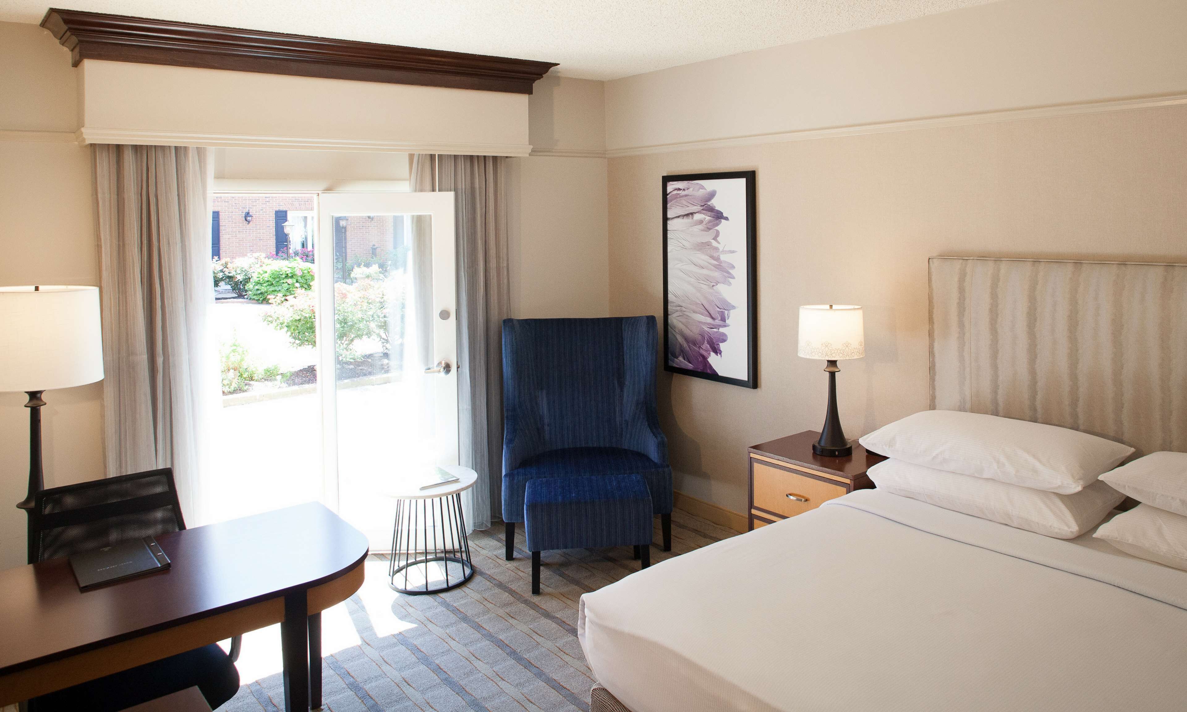 Une chambre d’hôtel comprenant un lit, deux tables de chevet avec des lampes, une chaise, une petite table ronde et une fenêtre avec des rideaux.