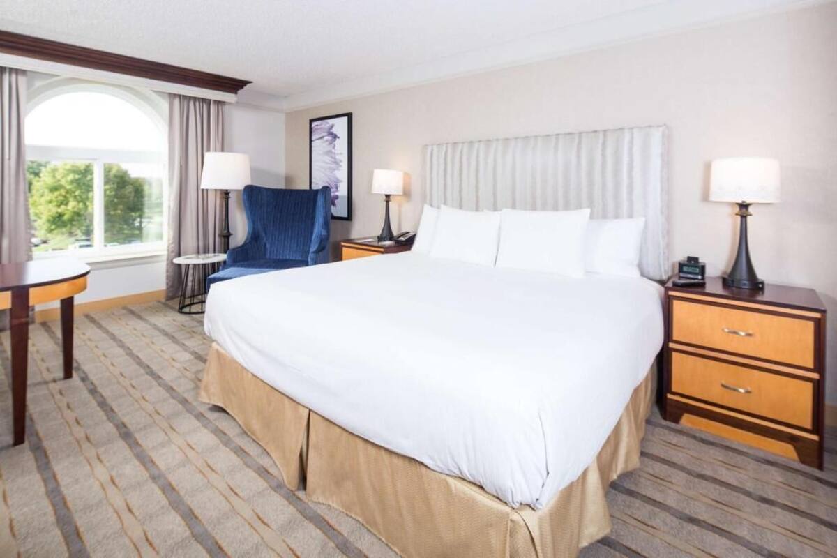 Een hotelkamer met een groot bed, twee nachtkastjes met lampen, een stoel, een kleine tafel en een raam met gordijnen.