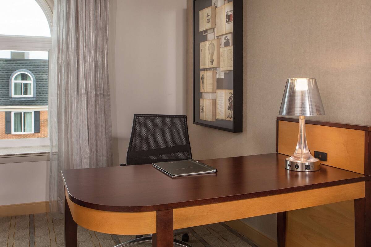 Een hotelkamer met een houten bureau, een laptop, een lamp, een lijst met foto’s aan de muur en een raam met uitzicht op een gebouw.