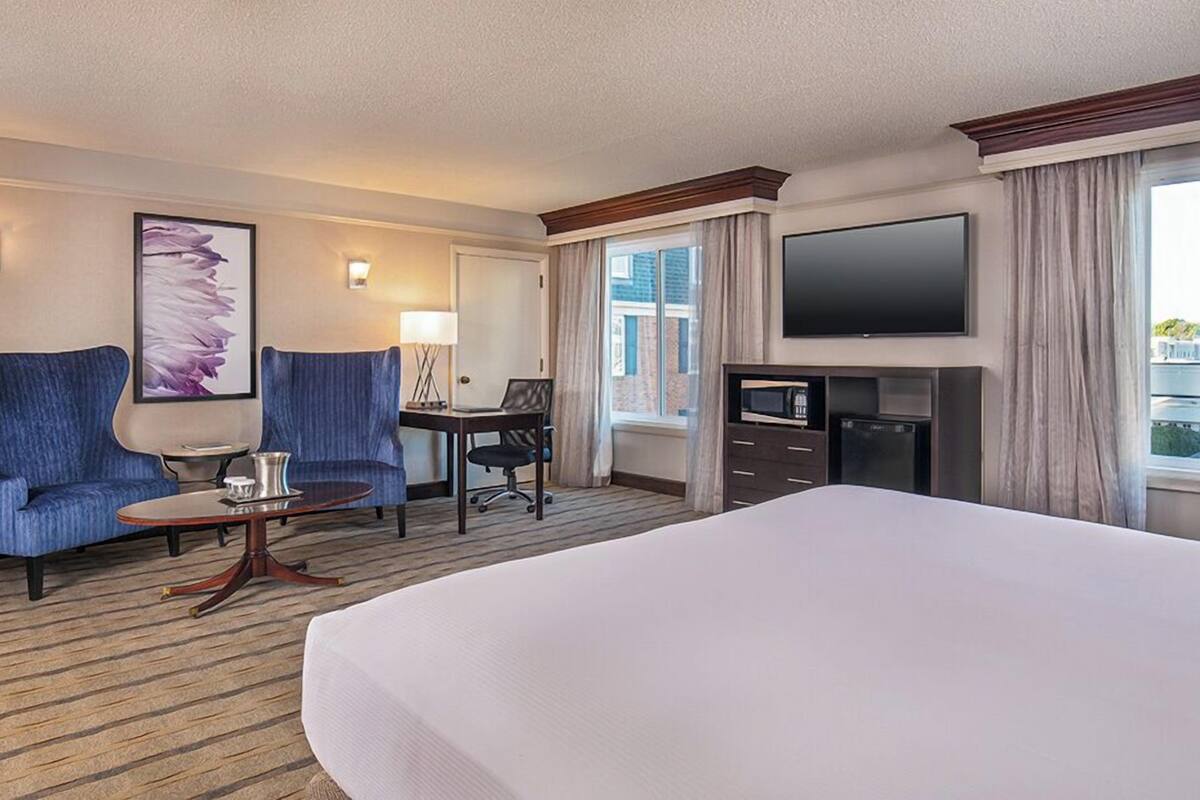 Een hotelkamer met een groot bed, twee fauteuils, een bureau met stoel, een flatscreen televisie en een raam met gordijnen.